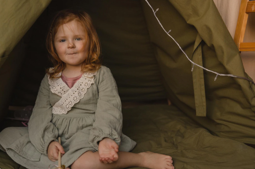 Little redhaired caucasian baby girl wear dress looks at camera while sitting in green tent indoors children lifestyle concept