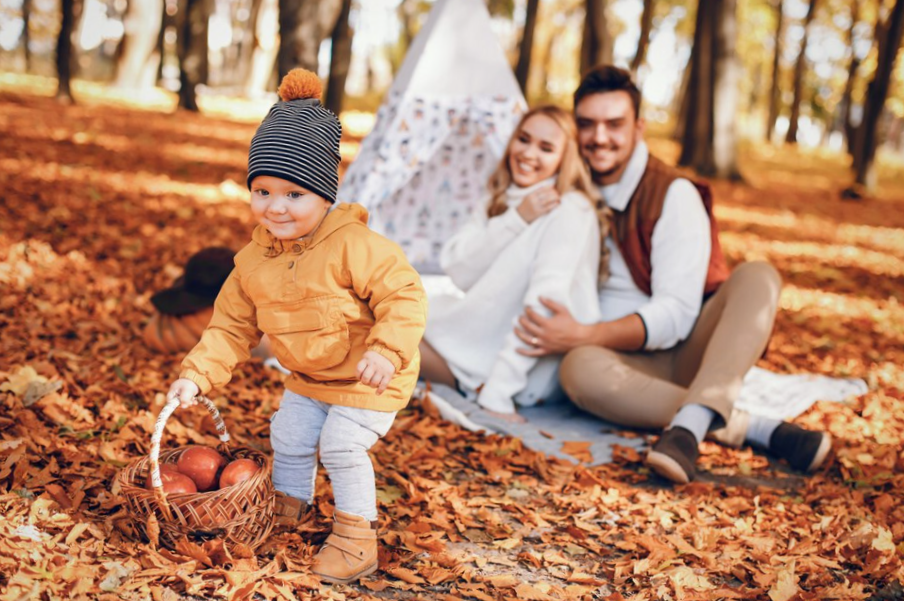 smiling baby wearing a wind proof jacket holding a basket with apples and his parents in the background 