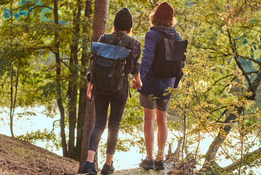 Romantic couple is standing near river in green bright forest and looking foward. they have backpacks and hats.