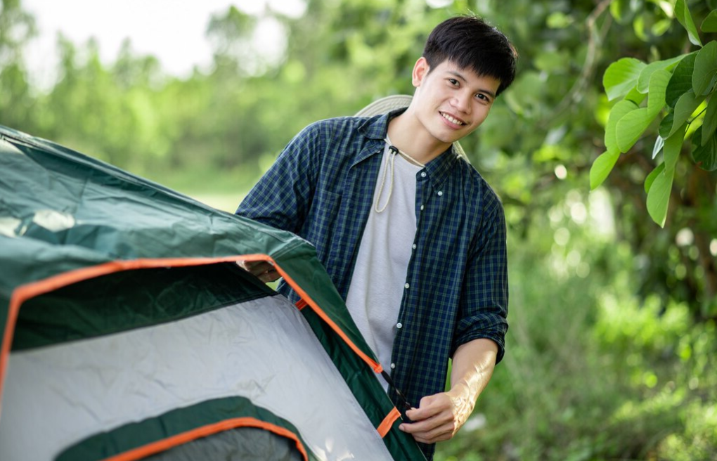 Smiley young traveler man setting a tent on the camping in the forest on summer vacation